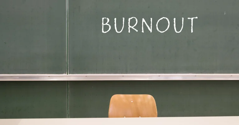 A teacher’s empty desk chair sits in front of a blackboard with the word “BURNOUT” written on it in chalk.