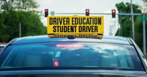 A yellow sign indicating a student driver is behind the wheel sits atop a car that is stopped at a red light.