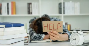 A student rests her head on her desk, feeling overwhelmed, and holds up a sign that says “HELP”.