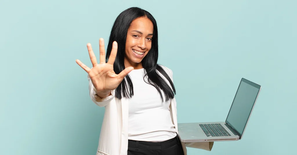 A young teacher in a white blazer holds up the number five with her right hand while holding a computer with her left hand.