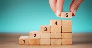 A hand stacks steps of wooden blocks numbered 1-5.