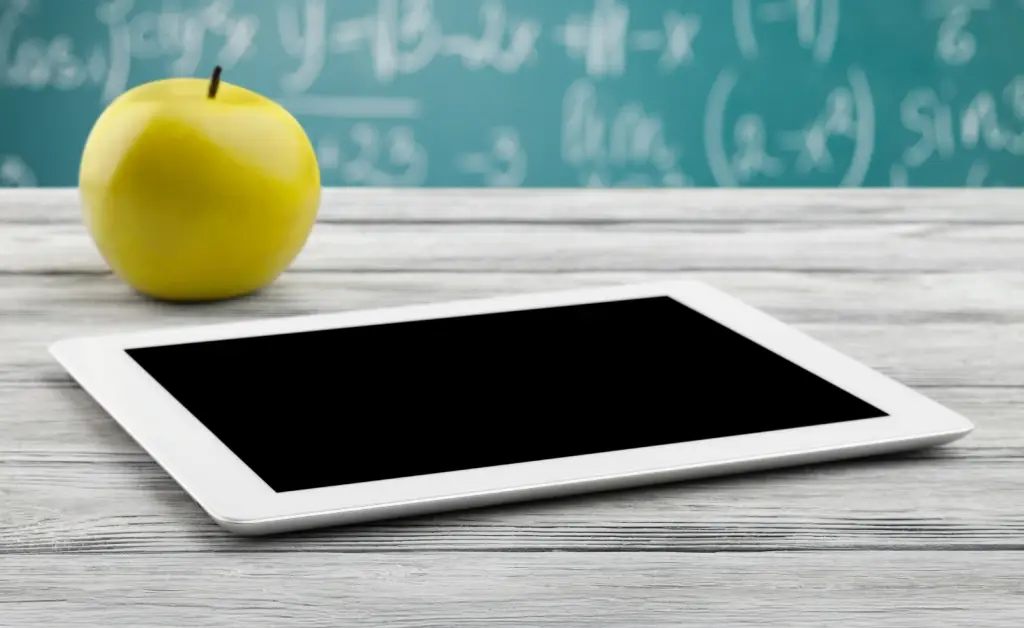 A yellow apple sits on a table next to a tablet in front of a blackboard covered in math problems