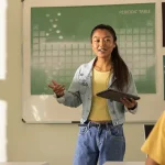 A student stands in front of her class giving a presentation while her teacher looks on with pride.