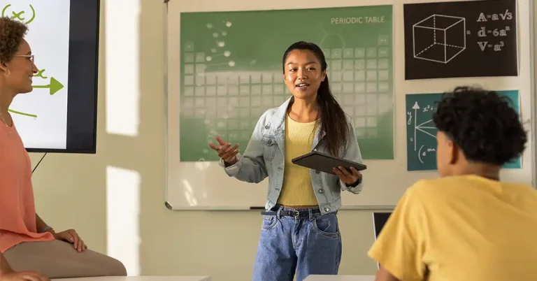 A student stands in front of her class giving a presentation while her teacher looks on with pride.