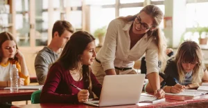 An AP teacher leans over at her student’s desk, assisting her with coursework on a laptop.